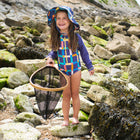 A smiling girl stands on a rocky beach wearing a 'UV Protective Long Sleeve Swimsuit Multi Check' and matching hat, holding a fishing net.